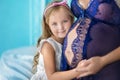 Beautifull young brunette pregnant woman in awesome purple dress close to blue sofa and cute flowers together with daughter. Royalty Free Stock Photo