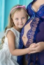 Beautifull young brunette pregnant woman in awesome purple dress close to blue sofa and cute flowers together with daughter. Royalty Free Stock Photo