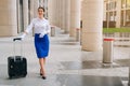 A beautifully smiling flight attendant with red lipstick in white gloves stands next to a suitcase. Girl carries luggage Royalty Free Stock Photo