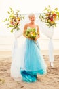 Beautuful blonde short haired woman posing in blue tulle long train dress near wedding flower arch with tropical bouquet Royalty Free Stock Photo