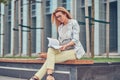 Charming blonde female in modern clothes, studying with a book, sitting on a bench in the park against a skyscraper. Royalty Free Stock Photo