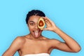 Beauty Concept. Young african woman isolated on blue covering eye with avocado showing tongue smiling playful Royalty Free Stock Photo