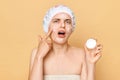 Beauty facial treatment. Shocked displeased woman wrapping in towel with shower cap on head cream in her hands having problems