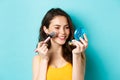 Beauty. Modern girl with candid smile applying make up with brush, looking in pocket mirror, standing over blue