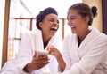 Beauty, phone and spa with women laughing in robes for luxury pampering or treatment together. Happy, app and social