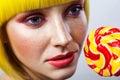 Beauty portrait of calm cute young female model with freckles, red makeup and yellow wig, holding colorful candy stick near face
