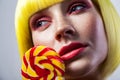 Beauty portrait of calm cute young female model with freckles, red makeup and yellow wig, holding colorful candy stick near face Royalty Free Stock Photo