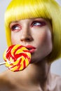 Beauty portrait of cute young fashion model with freckles, red makeup, yellow wig, holding colorful candy stick on lips and Royalty Free Stock Photo