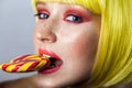 Beauty portrait of cute young female model with freckles, red makeup and yellow wig, holding and bitting her colorful candy stick