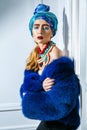 Beauty portrait of fashion model with colored headwear, blue fur coat red eyebrow and lips makeup and necklace. Royalty Free Stock Photo