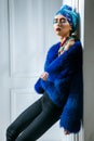 Beauty portrait of fashion model with colored headwear, blue fur coat red eyebrow and lips makeup and necklace. Royalty Free Stock Photo
