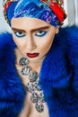 Beauty portrait of fashion model with colored headwear, blue fur coat red eyebrow and lips makeup and necklace. Royalty Free Stock Photo