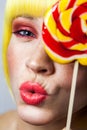 Beauty portrait of funny cute young female model with freckles, red makeup and yellow wig, holding colorful candy stick, kissing Royalty Free Stock Photo