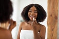 Beauty portrait of pretty young black woman applying lipstick and looking at mirror indoors. Organic makeup products