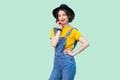 Beauty portrait of pretty young hipster girl in blue denim overalls, yellow shirt, black hat standing with hand on waist and Royalty Free Stock Photo
