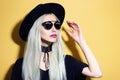 Beauty portrait of young fashion blonde hipster girl with pink lips, wearing sunglasses, black hat and choker, isolated on yellow. Royalty Free Stock Photo