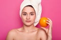 Beauty portrait of young girl with nacked shoulders. Lady holds orange, has calm facial expression. Charming female with towel