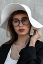 Beauty portrait young woman with clean skin in vintage glasses in black fashion jacket in straw white elegant summer hat near gray Royalty Free Stock Photo