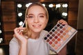 Beauty room. Cheerful nice young woman look on camera and smile. She applying eyeshadows with brush. Colorful palette in Royalty Free Stock Photo