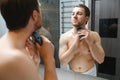 beauty, shaving, grooming and people concept - close up of young man looking to mirror and shaving beard with trimmer or Royalty Free Stock Photo