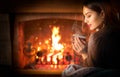 Beauty young Christmas woman sitting near fireplace in dark room at home and drinking hot beverage from a big cup