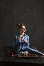 Beautyful young woman drinks coffee on black background in studio. Multi-colored cupcakes on black tray. Vertical frame Royalty Free Stock Photo