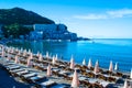 Sun loungers and umbrellas on public beach at summer mornin. Rafailovici buildings on background. Calm sea. Blue sky