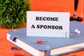 BECOME A SPONSOR text written on a business card standing with a clip on a diary, a notebook on an orange background