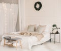 Bed with beige knitted plaid and a cup. Interior of stylish cozy bedroom with wreath on the wall. Christmas, New year home decor