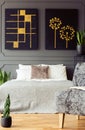 Bed with pillows under black and gold posters in bedroom interior with plants and chair. Real photo