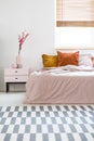 Bed with pink sheets and orange cushions standing in white bedroom interior with carpet, window with wooden blinds and bedside ta