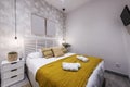 Bedroom with a double bed with a wooden Venetian-style headboard with a mustard-colored blanket, white bedside tables with drawers