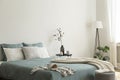 Bedroom interior with sage green and white sheets and cushions and a blanket. Black metal table with vases beside the bed. A lamp