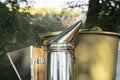 Beekeeper Smoker smokes white smoke. Apiary background. Beekeeping tool close up. Smoker stands on the ground on the green grass.