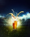 Beer and foam splashes. Mug with lager beer standing on grass at football stadium over evening sky with flashlights