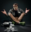 Beggar, pauper man in jacket, jeans and shoes sits cross legged on floor by upside down hat full of dollars cash