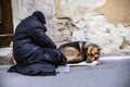 A beggar woman asks for alms with her dog. The dog laying next to the old woman