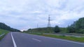 Behind a paved highway with metal fencing and markings, a high-voltage power line runs on a hill among a cornfield. On a summer ev