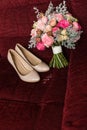Beige bridal shoes and a wedding rings lying on a red armchair. Wedding bouquet with purple and pink roses out of focus. Royalty Free Stock Photo