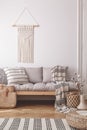 Beige, handmade macrame on a white wall above a couch with a striped linen pillow in a natural living room interior