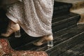 Beige shoes with a transparent heel on the bride`s legs go along the wooden staircase, evening dress with embroidery Royalty Free Stock Photo