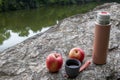 Beige thermos bottle, cup of tea or coffee, and red apple on rock over the river. Beautiful summer, spring, autumn landscape. Royalty Free Stock Photo