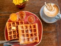 Belgian waffles and cappuccino served on a rustic wooden table with fresh a vase of fresh flowers.