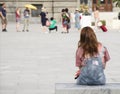 One young teenage girl with ginger hair in jeans overall sitting and waiting on city bench on town square, from behind