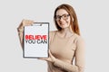 Believe you can. Beautiful young business woman wearing glasses holds a clipboard with mock up space isolated on gray background Royalty Free Stock Photo