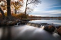 Smooth milky stream of water cascading down rocks from a glassy still lake surrounded by fall foliage.