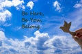 Belong symbol. Man hand holding wooden bird on cloud blue sky background. Words `be here, be you, belong`. Business, belonging a