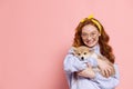 Cheerful redhead young girl with long curly hair holding cute little puppy of corgi dog isolated on pink background