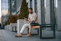 On the bench with smartphone and bag. Handsome black man is outdoors near the business building Royalty Free Stock Photo