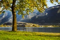 A bench under the tree next to the lake among the mountains in the sunrising light. Royalty Free Stock Photo